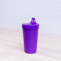 A REPLAY Re-Play No-Spill Sippy Cup in purple, featuring a spill-proof lid, is placed on a light wooden surface. Made from FDA-approved recycled HDPE plastic, this sustainable cup sits against a backdrop of a white tiled wall.