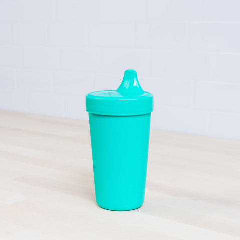 A Re-Play No-Spill Sippy Cup from REPLAY, featuring a bright teal color and spout lid, sits on a light wooden surface against a white tile background. Crafted from FDA-approved recycled plastic, the simplistic design is ideal for young children transitioning from bottles to regular cups.