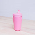 The Re-Play No-Spill Sippy Cup, a pink, FDA approved recycled plastic sippy cup with a spout lid from the brand REPLAY, is placed on a light-colored wooden surface. The background consists of white tiled walls.