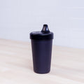 A black, textured Re-Play No-Spill Sippy Cup from REPLAY, made from FDA approved recycled plastic, sits on a light wooden surface against a white tiled background.