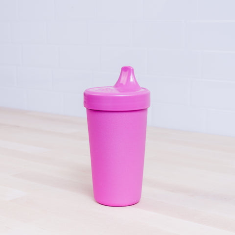 A pink REPLAY Re-Play No-Spill Sippy Cup with a spout lid sits on a light wooden surface against a white tile background. Made of FDA-approved recycled plastic, the cup boasts a matte finish.
