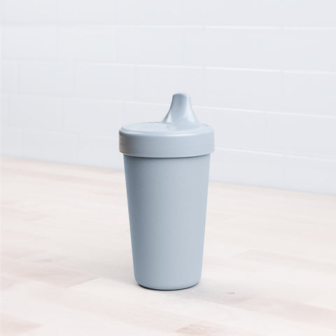 A Re-Play No-Spill Sippy Cup by REPLAY, light gray in color and featuring a spouted lid made from FDA-approved recycled plastic, sits on a light wooden surface. The background showcases white subway tiles.