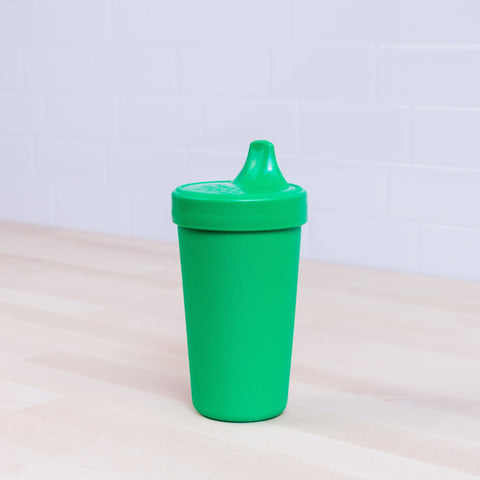 The Re-Play No-Spill Sippy Cup from REPLAY, made from FDA-approved recycled plastic with a spout lid, sits on a light wooden surface. The background features a white tiled wall.