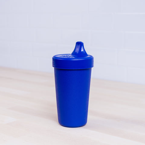 A Re-Play No-Spill Sippy Cup from REPLAY, crafted from FDA-approved recycled plastic, sits on a light wooden surface. The blue plastic cup features a convenient spout lid, while the clean and minimalistic background is set off by a white tiled wall.