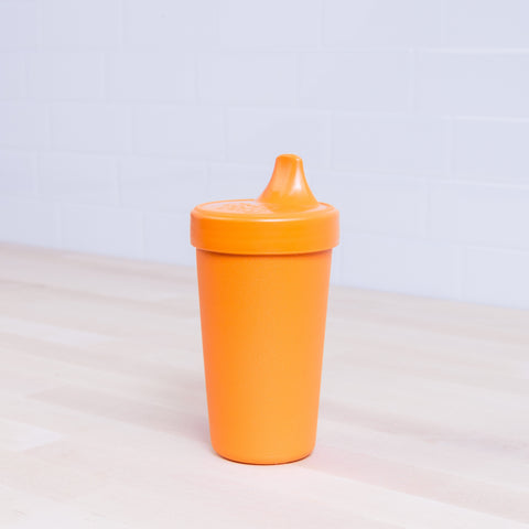 A Re-Play No-Spill Sippy Cup from REPLAY, featuring an orange spout lid, is placed on a light wooden surface against a white tiled wall. Made from FDA-approved recycled plastic, it's both practical and eco-friendly.