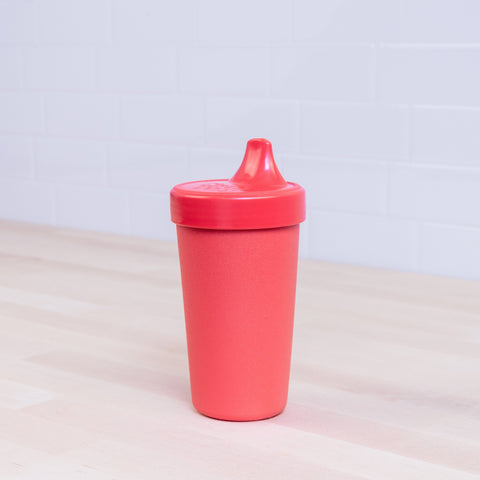 A Re-Play No-Spill Sippy Cup in red, featuring a spout lid and crafted from FDA-approved recycled plastic by REPLAY, is placed on a light wooden surface. The background consists of white tiled walls.