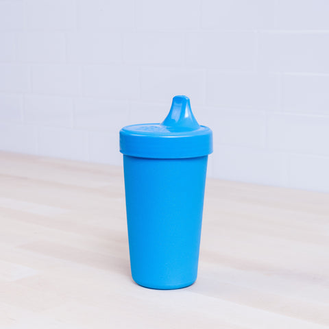 A blue Re-Play No-Spill Sippy Cup, crafted from FDA approved recycled plastic and featuring a spout lid, is placed on a light wooden surface. The background showcases white subway tiles.