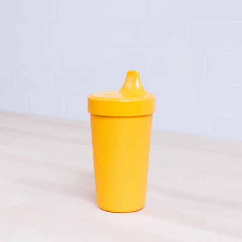 The Re-Play No-Spill Sippy Cup by REPLAY, a vibrant yellow sustainable HDPE cup, sits on a light wooden surface with a white tiled background. This cylindrical sippy cup features a spout lid and has a textured surface for easy gripping. It is made from FDA approved recycled plastic.