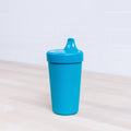 A blue Re-Play No-Spill Sippy Cup from REPLAY, featuring a matching lid, sits on a light-colored wooden surface. Crafted from FDA-approved recycled plastic HDPE, the cup is upright with its spout closed. The white tiled wall in the background highlights its sustainable design.