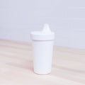 A white Re-Play No-Spill Sippy Cup from REPLAY, made from FDA approved recycled plastic, sits on a light wooden surface. Against a white tiled wall backdrop, the cup features a spout lid and retains a simple design with no visible patterns.