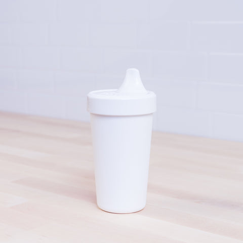 A white Re-Play No-Spill Sippy Cup from REPLAY, made from FDA approved recycled plastic, sits on a light wooden surface. Against a white tiled wall backdrop, the cup features a spout lid and retains a simple design with no visible patterns.