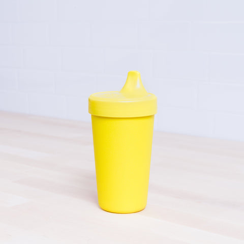 A Re-Play No-Spill Sippy Cup by REPLAY, featuring a bright yellow, textured design and a spill-proof lid with a spout on top, rests on a light wooden surface. The background showcases light-colored, tiled walls. This sippy cup is made from FDA-approved recycled plastic.