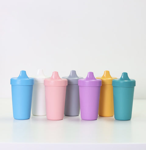 Seven Re-Play No-Spill Sippy Cups from the REPLAY brand, crafted from FDA-approved recycled plastic, are displayed in a row on a white surface. The cups showcase an array of colors: blue, pink, white, gray, purple, yellow, and teal. The background is plain white.