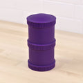 A durable cylindrical purple REPLAY Re-Play Snack Stack with grooved edges and three stacked sections stands upright on a light wooden surface. Made from recycled material, it is set against a background of white tiled walls.