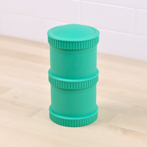 A Re-Play Snack Stack from REPLAY, a teal-colored, cylindrical-shaped plastic container with ribbed edges and a lid crafted from recycled material, is placed on a light-colored wooden surface in front of a white tiled wall.