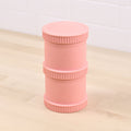 A Re-Play Snack Stack by REPLAY, featuring a pink cylindrical design made from recycled material with ribbed sections and the Rastaclat logo embossed on the side, is placed upright on a light wooden surface with a white tiled wall in the background.