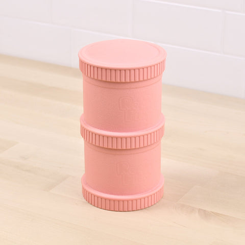 A Re-Play Snack Stack by REPLAY, featuring a pink cylindrical design made from recycled material with ribbed sections and the Rastaclat logo embossed on the side, is placed upright on a light wooden surface with a white tiled wall in the background.