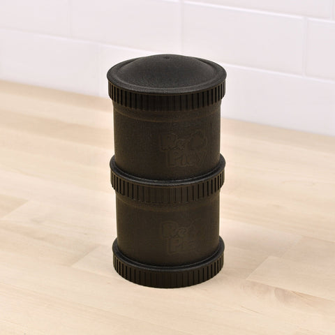 A cylindrical, black Re-Play Snack Stack from REPLAY features indented grooves and the text "Red Play" embossed on it. Made from recycled material, this durable snack container is segmented into three parts and sits on a light wooden surface in front of a white tiled wall.