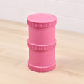 A pink Re-Play Snack Stack made by REPLAY, crafted from recycled material with three distinct segmented sections, stands vertically on a light wooden floor against a white tiled wall. The stack has ribbed edges on the top and bottom, with the words "Rock Fluo" embossed on its side.