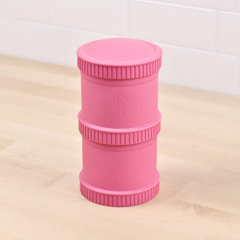 A pink Re-Play Snack Stack made by REPLAY, crafted from recycled material with three distinct segmented sections, stands vertically on a light wooden floor against a white tiled wall. The stack has ribbed edges on the top and bottom, with the words "Rock Fluo" embossed on its side.