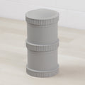 The Re-Play Snack Stack by REPLAY is a gray cylindrical container with two segmented sections, each featuring ridged edges. Crafted from recycled material, this piece of sustainable tableware stands upright on a light wooden surface with a white tiled wall in the background.