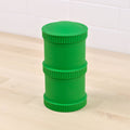 A green, cylindrical plastic Re-Play Snack Stack from REPLAY, made from recycled materials, featuring three stacked sections with ribbed sides for easy grip, is displayed on a light wooden floor against a white tiled wall – ideal for durable snack storage.