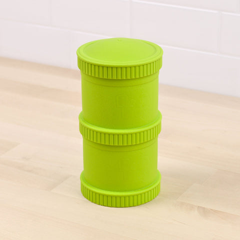 The REPLAY Re-Play Snack Stack is a lime green, cylindrical container featuring a textured lid and base, crafted from recycled material. Its innovative design includes a narrower central section and ring-like grooves around the top, middle, and bottom areas. This eco-friendly addition to your sustainable tableware collection is showcased on a light wood surface with a white tiled backsplash.