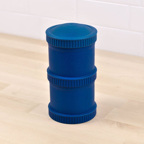 The REPLAY Re-Play Snack Stack, a large blue cylindrical container crafted from recycled material and featuring textured rings at the middle as well as the top and bottom, stands on a light wooden floor against a white tiled wall.