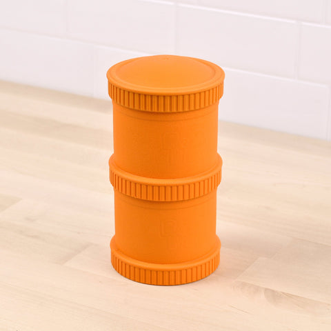 A Re-Play Snack Stack, an orange cylindrical object made from recycled material with ribbed edges, stands upright on a light wooden surface. The snack stack has two thick bands around its middle section and appears to be made of plastic. The background features light-colored tiled walls.