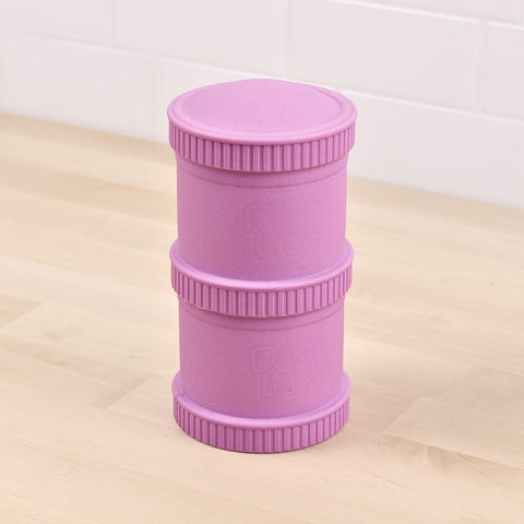 A *REPLAY* Re-Play Snack Stack, a cylindrical object colored pinkish-purple and featuring textured bands near the top, middle, and bottom that resemble a durable snack stack or storage unit, is placed on a light wooden surface with a tiled background.