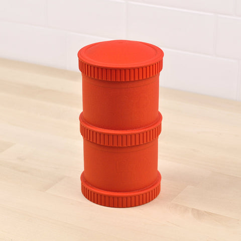 A red, cylindrical dog toy from the "Re-Play Snack Stack" by REPLAY, made from recycled material with two textured bands, is placed on a wooden floor against a white tiled wall.