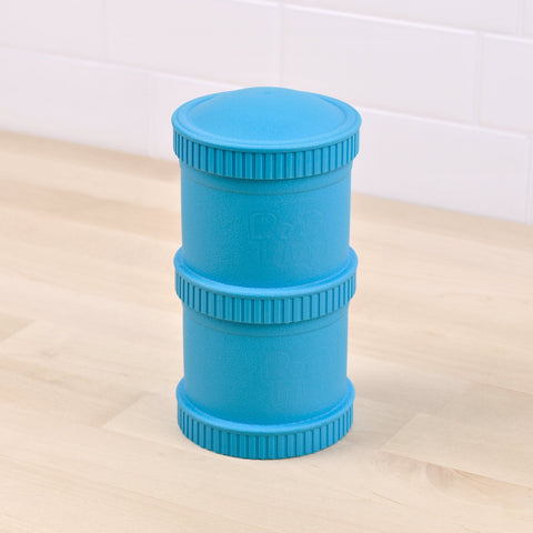 The Re-Play Snack Stack, a blue cylindrical dog puzzle toy with ridged edges and grooves crafted from recycled material by REPLAY, stands on a light wood floor in front of a white tiled wall. The toy, embossed with the REPLAY logo, features alternating sections designed to hold treats.