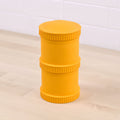 The Re-Play Snack Stack by REPLAY is a yellow, cylindrical container made from recycled material, boasting a textured surface and ribbed edges. It stands on a light-colored wooden surface and features two seamlessly stacked sections with a uniform appearance. In the background, there is a white tiled wall.