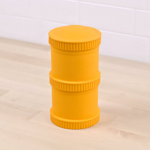 The Re-Play Snack Stack by REPLAY is a yellow, cylindrical container made from recycled material, boasting a textured surface and ribbed edges. It stands on a light-colored wooden surface and features two seamlessly stacked sections with a uniform appearance. In the background, there is a white tiled wall.