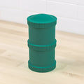 A green Re-Play Snack Stack from REPLAY, featuring two ridged sections, sits on a light wooden surface. Made from recycled material, it resembles durable snack stacks or storage jars. White tile walls are visible in the background.