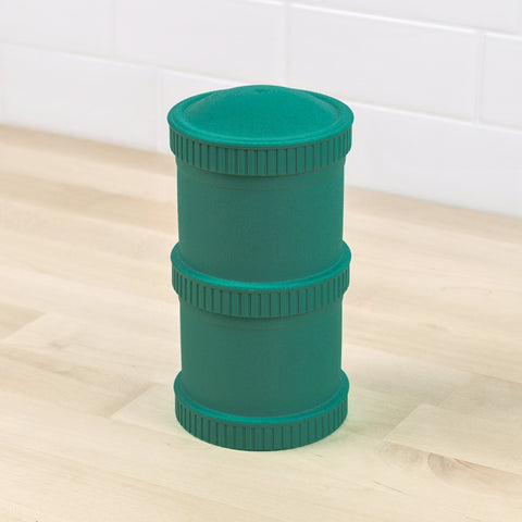 A green Re-Play Snack Stack from REPLAY, featuring two ridged sections, sits on a light wooden surface. Made from recycled material, it resembles durable snack stacks or storage jars. White tile walls are visible in the background.