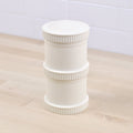 A white cylindrical Re-Play Snack Stack from REPLAY with ribbed edges stands on a light wooden surface against a tiled white background. Crafted from recycled materials, the container is segmented into three parts with "RICE PLAY" embossed on the middle section. This piece exemplifies sustainable tableware at its finest.