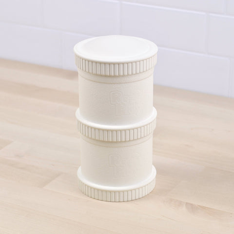 A white cylindrical Re-Play Snack Stack from REPLAY with ribbed edges stands on a light wooden surface against a tiled white background. Crafted from recycled materials, the container is segmented into three parts with "RICE PLAY" embossed on the middle section. This piece exemplifies sustainable tableware at its finest.