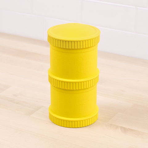 A large, yellow cylindrical container with ribbed edges, resembling stacked rings or sections. Made of recycled material, the durable REPLAY Re-Play Snack Stack is placed on a light wood surface with a white tile wall in the background.