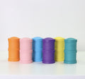 Re-Play Snack Stacks from REPLAY, made from recycled materials, are lined up in a row on a white surface. These durable cylindrical containers come in six distinct colors: orange, pink, blue, purple, yellow, and teal. Each container features a ribbed texture and a rounded top.