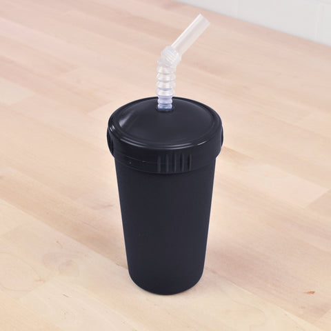 The Re-Play Straw Cup by REPLAY, a black, reusable tumbler with a black lid and a clear, flexible straw, sits on a wooden surface. Made in the USA from sustainable materials, this matte tumbler features a recycled straw inserted through a hole in the center of the lid.