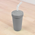 A light-colored wooden surface holds a REPLAY Re-Play Straw Cup, which is gray, insulated, and equipped with a screw-on lid. The cup includes a re-usable straw made from recycled plastic, featuring a flexible, ribbed segment near the top for easy bending—emphasizing its sustainable design.