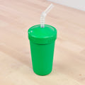 A Re-Play Straw Cup in green, featuring a screw-on lid and a flexible, translucent straw, is placed on a light wooden surface. The sustainable cup from REPLAY, made in the USA, stands upright with the straw slightly bent. The background is plain and uncluttered.