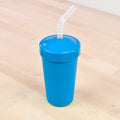 A Re-Play Straw Cup by REPLAY, featuring a vibrant blue body and a coordinating lid, sits on a light wooden surface. The lid includes a clear plastic spiral straw extending from the top. Made in the USA, this eco-friendly cup is ideal for cold beverages.