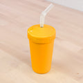 An REPLAY Re-Play Straw Cup, crafted from bright orange, reusable plastic and featuring a tightly fitting lid along with a flexible, transparent straw made from recycled materials, stands upright on a light wood surface. It is the only object in the image, set against a plain and unobtrusive background.