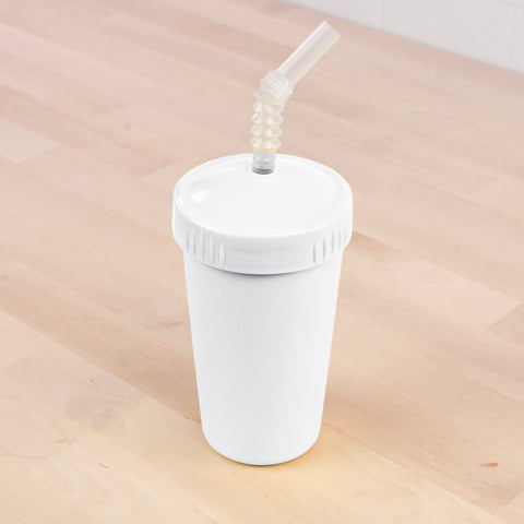 A white plastic Re-Play Straw Cup from REPLAY with a lid and a flexible straw sits on a light wooden surface. The cup features a textured screw-on lid, and the straw is inserted through a small hole in the center of the lid. This sustainable design reflects its commitment to eco-friendly practices, proudly made in the USA.