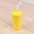 The REPLAY Re-Play Straw Cup, a vibrant yellow textured tumbler made in the USA, sits on a light wooden surface with a matching yellow lid. The lid features an opening in the center through which a flexible, transparent recycled straw is inserted. The simple and unobtrusive background highlights this sustainable tumbler.