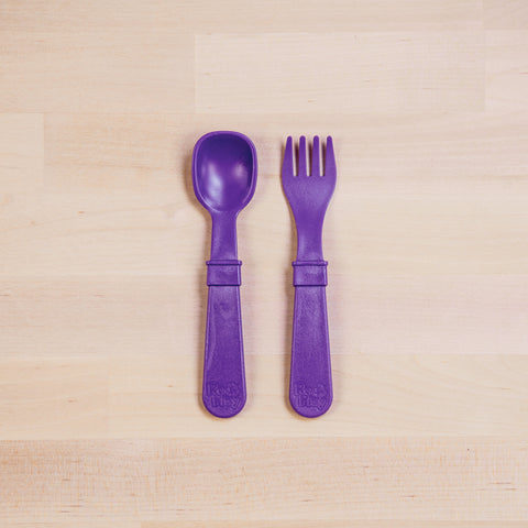 A set of toddler utensils from the REPLAY brand, consisting of a purple plastic spoon and fork made from recycled materials, placed side by side on a light wooden surface. The Re-Play Utensil Set features the spoon on the left and the broader-tined fork on the right. Both utensils have thick handles engraved with the "REPLAY" logo and are safe for dishwasher use.