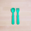 A Re-Play Utensil Set in bright turquoise, crafted from recycled plastic by REPLAY, lies side by side on a light wooden surface. The dishwasher-safe spork and spoon feature broad handles with raised sections in the middle; the spork boasts shallow tines while the spoon has a rounded bowl.