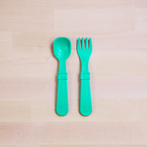 A Re-Play Utensil Set in bright turquoise, crafted from recycled plastic by REPLAY, lies side by side on a light wooden surface. The dishwasher-safe spork and spoon feature broad handles with raised sections in the middle; the spork boasts shallow tines while the spoon has a rounded bowl.